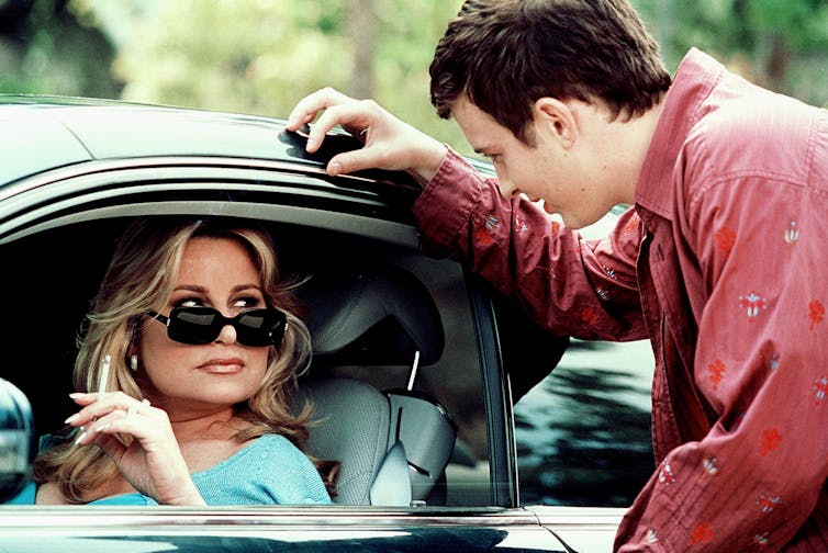 Jennifer Coolidge está sentada en el asiento de un automóvil, con gafas de sol y fumando por la ventanilla. La ventanilla está bajada para que pueda hablar con un adolescente.