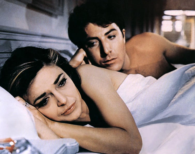 Dustin Hoffman y Anne Bancroft están acostados en la cama, después del coito. Ella está de espaldas a él y él está detrás de ella, mirándola inquisitivamente a la cara.